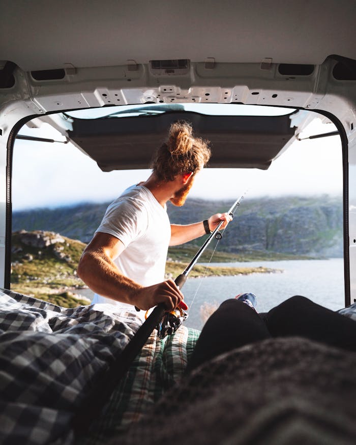 Man enjoys fishing from a van with a scenic lake view, embracing outdoor adventure and travel.