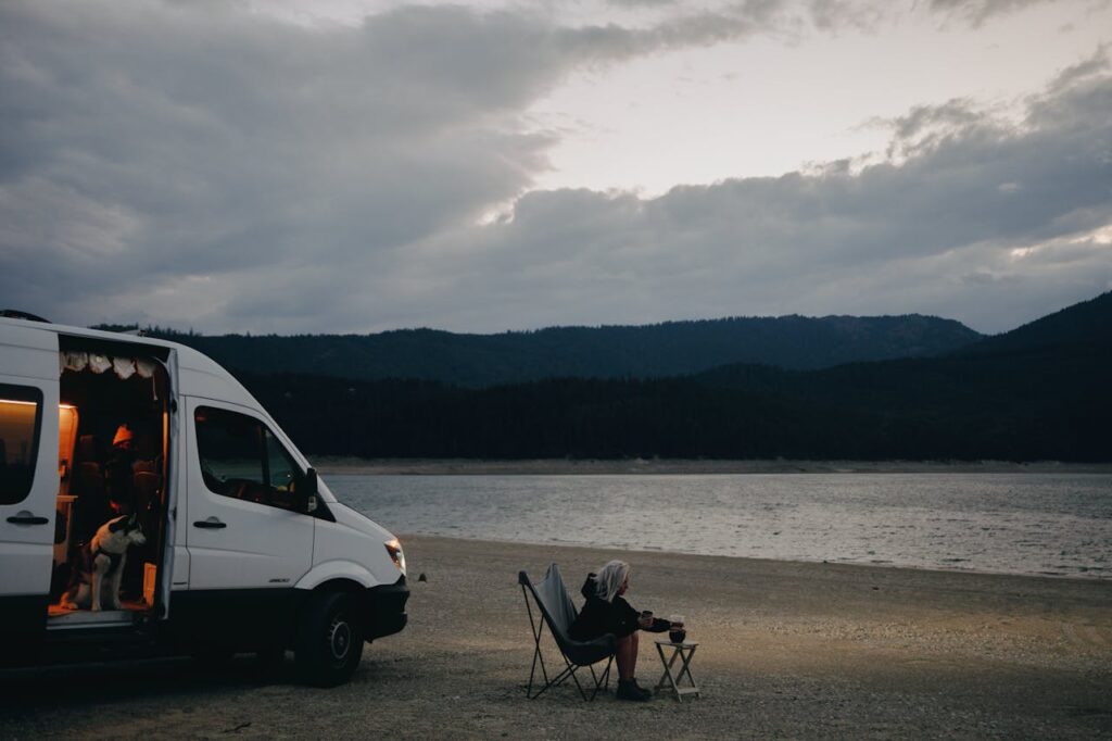 A woman relaxes by a lakeside, seated next to a campervan with a dog and folding chair under cloudy skies.
