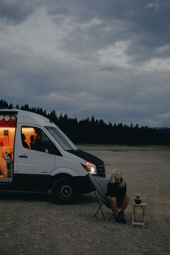An adult woman relaxes outside a campervan, sitting under cloudy skies with her dog by her side.