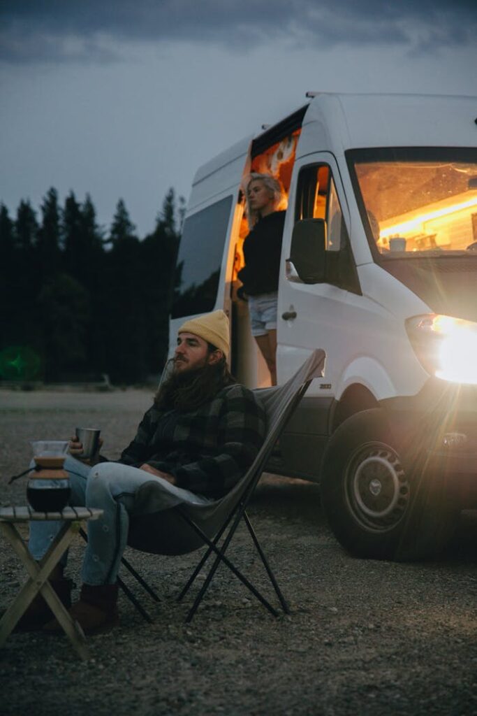 Bearded man in beanie relaxing with coffee by a lit camper van in the outdoors at dusk.